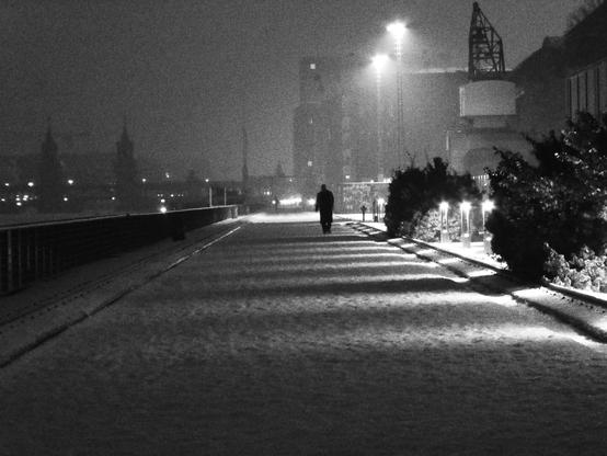 a very grainy black-and-white picture of a snowy path along the river. A person, only a black shape, is walking towards the camera. Some street lights high above are giving light to the scene.