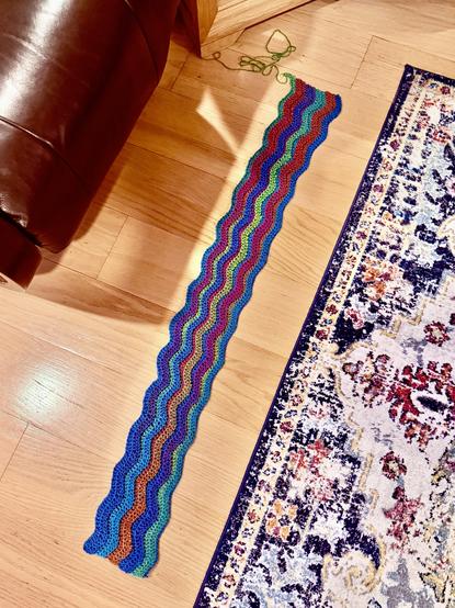 A long, narrow crochet ripple strip lies on a wooden floor between a leather sofa and a patterned rug. The wavy rows shift through blue, green, red, and orange, with a strand of green yarn still attached at one end.