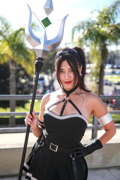 Cosplayer Joelynncosplay at Anime Los Angeles with the sun at her back.  She's dressed as Ubel from the anime Freiren.  Black dress with a black neck choker and straps running down her chest.  Black gloves and silver armband.  A silver trim along the top of the top of the corset like dress and a black belt with gold buckle.  She's holding the silver and black trident with a green magical crystal.