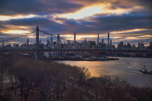 A panoramic view of a city skyline, featuring a prominent bridge connecting two sides of a river. The setting sun casts a colorful glow across the clouds, reflecting on the water, while bare trees are visible in the foreground. The scene captures the urban landscape at sunset.