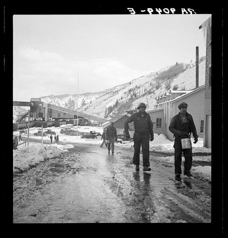 The image is a black and white photograph depicting an outdoor winter scene. It shows two individuals walking down a snowy street, with one person carrying containers on their shoulders which might be used for collecting water or other materials.

In the background, there's evidence of industrial activity such as large buildings that appear to be related to mining operations based on visible equipment and structures like conveyor belts and rail tracks filled with cars. The landscape is hilly, covered in snow, suggesting a cold climate likely associated with mountainous regions known for mining activities. 

The sky appears overcast, casting diffused light across the scene without creating harsh shadows or bright highlights.

Additional text overlaid at the top reads "3 - PHOP AT" which could be indicative of the photographer's notations on their negatives. The context and specific location are suggested to relate potentially to a mining town near Price, Utah known as Consumers based on provided references in the caption indicating its possible relation with Blue Blaze mine.

It’s worth noting that such photographs often serve historical purposes capturing daily life and work conditions during particular eras, providing insight into labor practices of miners.
