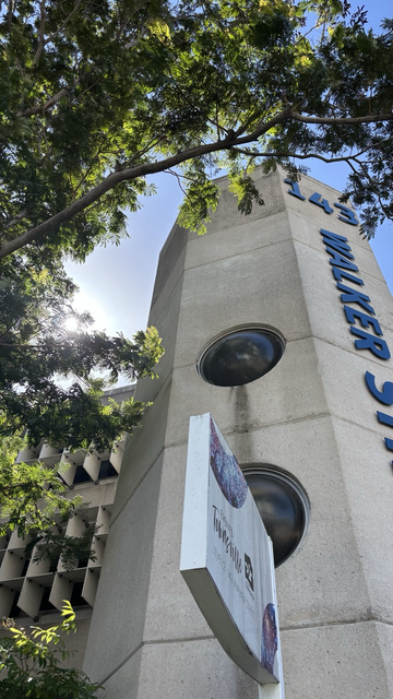 A hemispherical dark glass window punctures a brutalist concrete tower, the scene balanced by a lush green tree and the backdrop of a sunlit blue sky