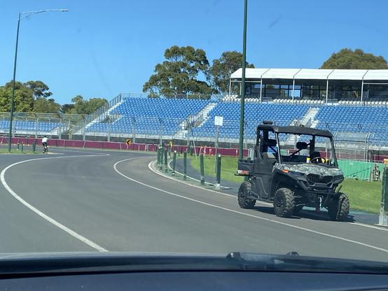 A black all-terrain vehicle is parked on a curved road near a racetrack. In the background, there are empty blue grandstands under a clear blue sky. A cyclist is riding on the track.