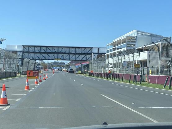 A view of a road leading to a racetrack, with construction activity evident. Traffic cones line the street, and signs indicate "Prepare to Stop." A pedestrian bridge crosses above, and a building structure is visible nearby. The sky is clear and blue