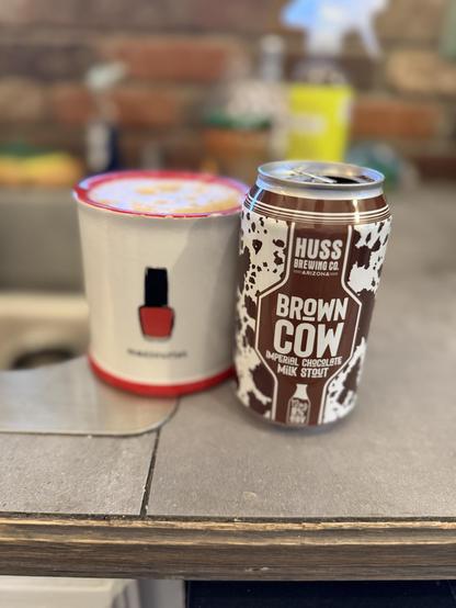 Some kind of coffee cup with a picture of a nail polish bottle is next to a brown and white can with a cowhide motif. This no’s not the right cup for this beer, but no one cares.