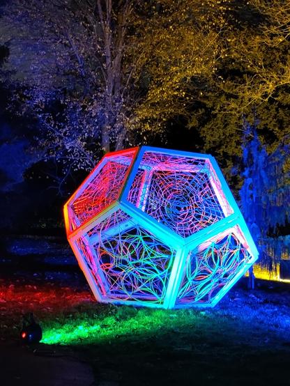 Eine Kunstinstallation in einem Park, im Hintergrund sind Bäume.
Das Objekt hat 12 Seiten die jeweils 5 Ecken haben. Die Flächen sind zusätzlich mit Mustern verziert.
Das Installation wird von verschiedenen Seiten bunt angestrahlt.