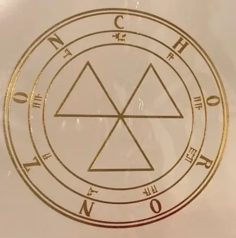 A close photo of a golden sigil over a beige surface with a glass-like reflective layer sitting on top of it, with some reflected image which is hard to be seen or distinguished. The sigil is specifically the sigil of Choronzon (an entity once described in Enochian magick and later by Aleister Crowley's Thelema). The central shape is quite similar to that of "radioactive hazard" (three equilateral triangular blades aligned to angles that are multiples of 120 degrees). Unknown origin (I found it on Google Images)