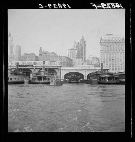 The image depicts a historical black-and-white photograph featuring several large ferries docked along the waterfront of what appears to be New York City. These boats are part of an integrated system for transporting passengers, likely connecting them with nearby train stations as indicated by signs reading "R.R.O." and other text on the structures above their docks.

In the background, a collection of prominent buildings rises against a gray sky. Among these edifices is one that stands out due to its pointed spire or turret-like architecture, which gives it an ornate appearance typical for late 19th-century urban skyscrapers and commercial landmarks in New York City's downtown area.

The water surface captures the movement of waves caused by either the passing ferries or a moderate current. The photograph has visible inscriptions at its top corner with alphanumeric codes such as "3-568P" that could be indicative of cataloging information, possibly related to historical archives where this photo might have been stored.

This image is evocative of early 20th-century urban transit systems and highlights a bygone era when river travel was integral for commuters in the bustling metropolis. The photograph seems like part of an archival collection that documents various aspects of city life during its heyday, providing historical context to transportation networks from over a c [...]