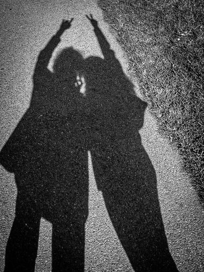 A monochrome photo of two people's long shadows on an asphalt path next to a grassy area. They are posing together with extended arms. One is doing the Sign of the Horns while the other does a peace sign.