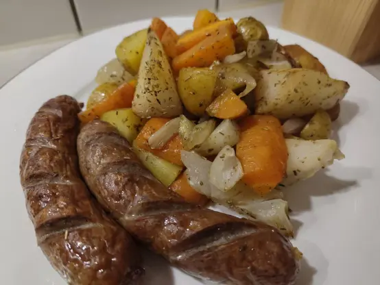 Zwei Bratwürste vom Wildschwein neben einem Haufem Ofengemüse aus Kartoffeln, Karotten, Fenchel und Birne auf einem Teller.