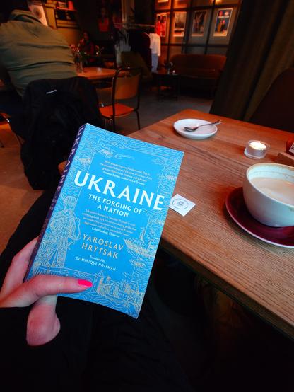Matt holds a blue book titled "Ukraine: The Forging of a Nation" by Yaroslav Hrytsak in a café setting, with a coffee cup and saucer on a wooden table.