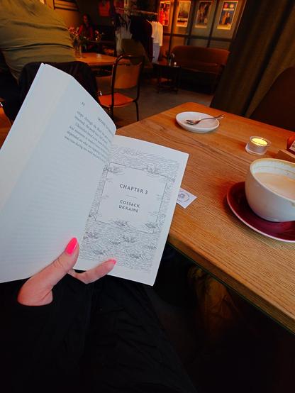 An open book showing Chapter 3 titled "Cossack Ukraine," held in a café with a coffee cup and saucer on a wooden table.