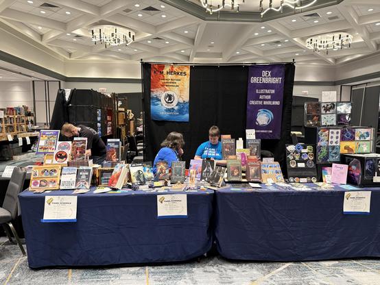 view of the Goshdarned Good Authors co-op table in the Capricon 46 Dealer room, with K M Herkes's books and bling on the left, Rhiannon Taylor's offering in t the center, and Dex Greenbright's books and bling on the right. Three people are visible behind the table,  heads bent over unseen items of interest. A tall black backdrop sits behind the table with brightly colored informational banners hung from the top.