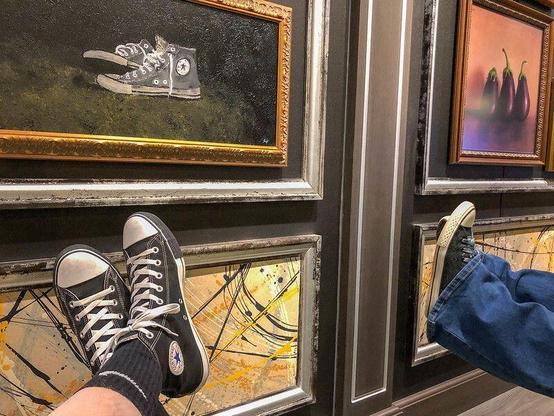 A wall of paintings, the most visible of which is a still life of a pair of classic black Chuck Taylor Converse high-top shoes. In front of the painting are the feet of my wife and I, both wearing Converse shoes; mine match the pair in the painting.