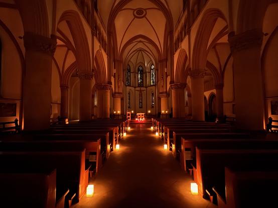 Der dunkle Innenraum einer Kirche mit hohen gotischen Spitzbögen und hölzernen Kirchenbänken. Der Mittelgang ist durch kleine, warm leuchtende Kerzen auf dem Boden gesäumt, die den Blick zum Altar führen, der im Hintergrund rötlich beleuchtet ist.