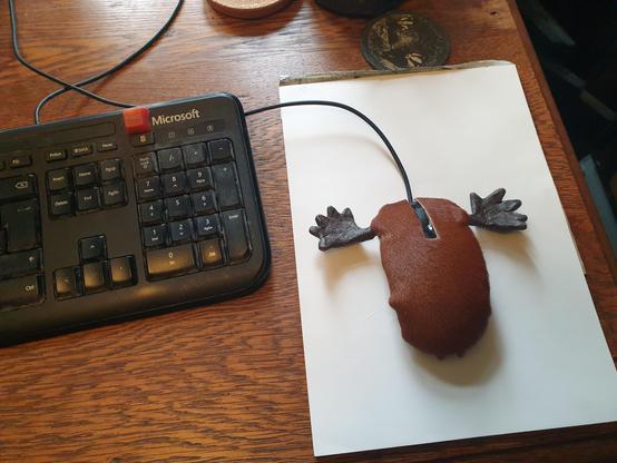 A computer mouse on a white pad next to a keyboard. It has been covered in fake, brown fur and had two modeling clay antlers glued to the sides next to the buttons