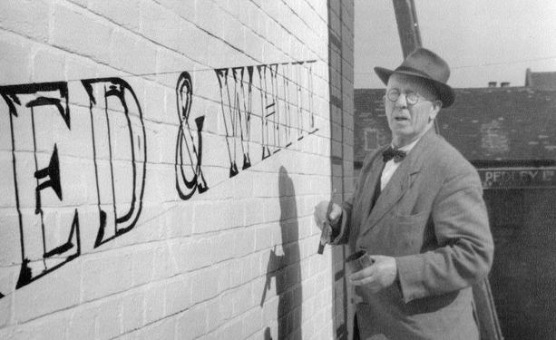 Black and white photo of a smartly dressed man posing with a brush and tin of paint in front of a whitewashed brick wall. He's been outlining letters on the wall in black, of which "...ED & WHITE" are visible on a diagonal.