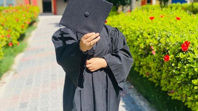 Naeema in graduation gown holding mortarboard in front of her face, standing on a paved walkway, with bushes and flowers on either side.