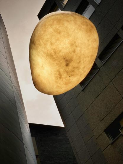 Eine übergroße, kugelförmige Leuchte hängt zwischen zwei modernen Gebäuden, die in einem warmen, goldenen Farbton vor einem bewölkten Himmel beleuchtet werden.