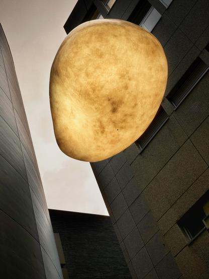 Eine übergroße, kugelförmige Leuchte hängt zwischen zwei modernen Gebäuden, die in einem warmen, goldenen Farbton vor einem bewölkten Himmel beleuchtet werden.