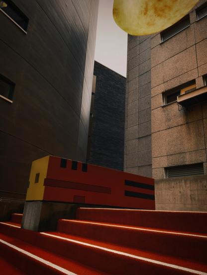 Eine urbane Szene mit stilisierten Stufen in leuchtendem Rot, die zu einer farbenfrohen, abstrakten Struktur führen. Die umgebenden Wände bestehen aus glatten und strukturierten Materialien, die durch weiche, natürliche Beleuchtung hervorgehoben werden. Ein großes kreisförmiges Objekt, das einem Mond ähnelt, ist oben sichtbar