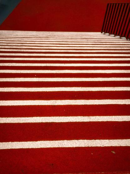 Ein Bild von Treppen mit abwechselnden roten und weißen Streifen, die nach unten führen. Ein schwarzes Geländer ist auf der rechten Seite sichtbar. Die Gesamtkomposition weist starke Kontraste in Farben und Mustern auf.