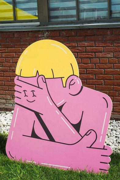 A colorful, cartoonish cutout of a person with a yellow hairstyle, playfully covering their face with one arm against a brick wall backdrop.