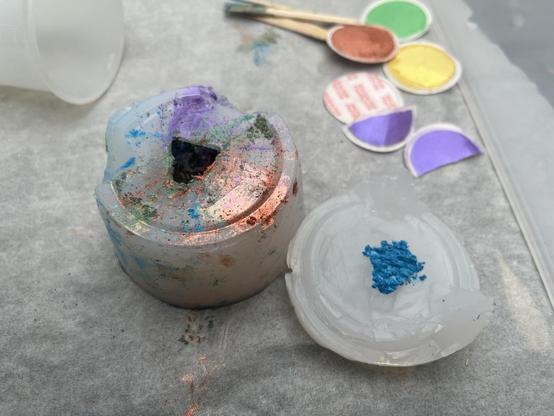 A frustum shaped silicone d20 mould with the cap off. The outside is stained with mica powder of the aforementioned colours, and the cap has a (too) large amount of cyan powder heaped on top of the face. 

Behind, there are coloured lids that have been peeled off the mica powder containers. There are also lollipop sticks with powder on the end.