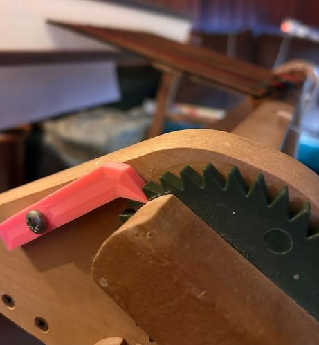 A close-up view of a mechanical component featuring a green gear and a pink 3D-printed lever, positioned on a wooden body. The background is slightly blurred, hinting at an indoor setting with some scattered items.