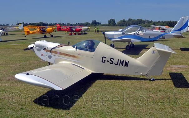 Side view of a small, single-engined, single-seat light aircraft, parked facing to the left on grass.
The plane is mostly cream coloured, with white engine cowling and #wingtips, and the black registration "G-SJMW" on the rear fuselage.
There is a gold stripe on the forward fuselage, with a small UK flag, and the black text "SD-1 Minisport".
Strong sunlight coming from the top left of the frame is casting a strong shadow on the ground under the plane.
Green grass extends into the distance, with several light aircraft parked on both sides of the image, facing in towards the middle.
Trees in the distance meet blue sky with a slight hint of haze.