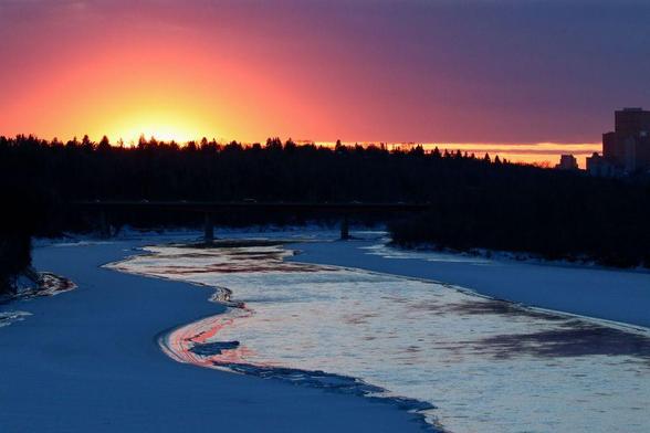 Photo of sunset over the partially frozen North Saskatchewan river. There is a bright orange and yellow burst where the sun is positioned, fading out to pinks and purples. The horizon is lined mostly with silhouetted trees, with some downtown buildings in silhouette on the right-hand side. A bridge crosses over the river with traffic just visible from the reflected sun light on the vehicles.