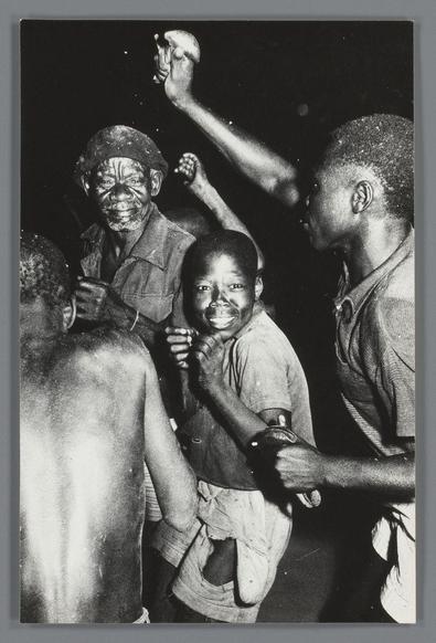 The image is a black and white photograph capturing the dynamic energy of several individuals during what appears to be an evening celebration or gathering. In the foreground, three young African-American boys are prominently featured: one on the left with his back partially turned towards us, another in the center smiling broadly at the camera, and the third on the right who is facing away but still visible from behind.

The boy in the middle stands out due to his joyful expression; he has wide eyes, an open mouth as if laughing or shouting, and hands clenched into fists. His skin shows some signs of wear or dirt, suggesting recent activity that could be related to play or work. The child on the right is similarly engaged but facing away from us, with a focus on his backside.

Behind them are two older men who seem to be partaking in the festivities as well. One man on the left wears glasses and has an intense expression while clenching his hand into a fist near his head level; he appears contemplative or perhaps cheering along. The other older man, standing behind him with slightly obscured features, also looks animated.

The background is dark and lacks distinct details, which puts emphasis on the figures in the foreground. There's no clear indication of where this event takes place—could be a street, an alleyway, or any public space conducive to gatherings—and it exud [...]
