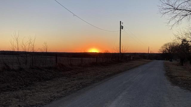 The orange-yellow sun just about to dip below the horizon.  A strip of orange at the horizon.   A gravel road goes away to straight west.  The sun is setting in the WSW.