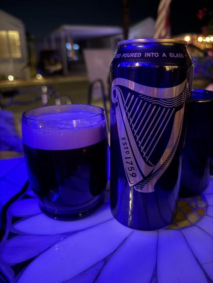 A black pint can with a gold harp logo is next to an undersized cup of beer with a thick tan head