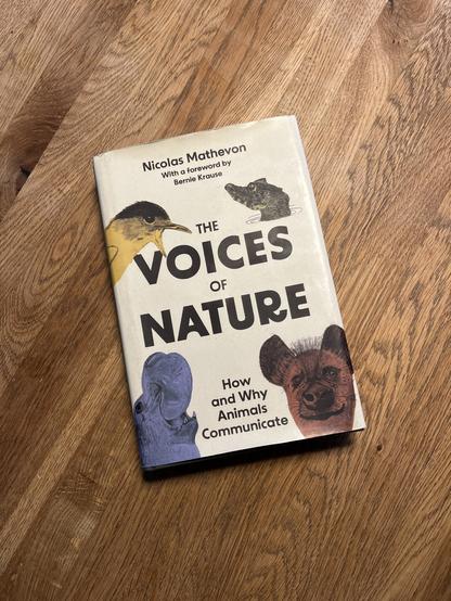 A book titled "The Voices of Nature - How and why animals communicate“ by Nicolas Mathevon, featuring illustrations of various animals such as a bird, a crocodile, a hippopotamus, and a hyena on the cover. 
The book is placed on a wooden surface.