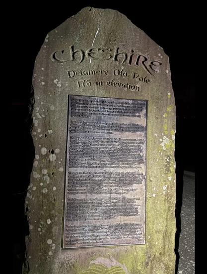 A stone monument with the word "Cheshire" carved at the top, featuring an inscription in English detailing historical information about the area, including its elevation of 176 meters. The stone is surrounded by a natural environment and is illuminated at night.