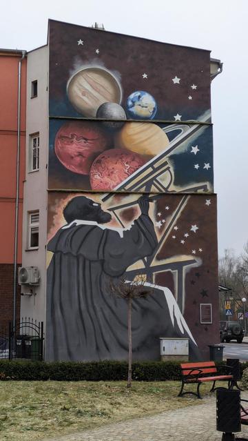 A mural showing an astronomer wearing a cloak, at a telescope. The person and its tools were depicted in dark gray, with the bearded head almost black, while its top edges were marked with white stripes. The person is then surrounded by a fiery outline. Above it, the planets of the solar system, in their usual colors, have been piled together in an unruly mass. The background is brown, with dark blue stripe in the middle. Over it all and to the right, a stripe is made of five-edged stars.