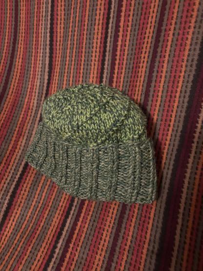 A green knitted watch hat, with a darker green ribbed brim and a paler crown that has a rotating pattern swirling upwards. It is sat on a striped brown/red/orange blanket.