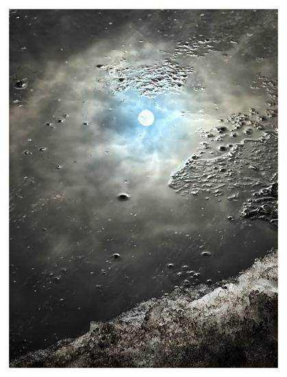 Closeup of a very shallow gray puddle on a city street. Little concrete bumps stick out of the water’s surface like islands, and a patch of dirty snow and ice slopes upward from left to right in the foreground at bottom frame. Reflected in the water is a pale winter sun, in a small opening of blue sky surrounded by gray clouds. Although it’s clear what the image is, it also makes me think of a snow-capped mountain lit by a waning sun, shining through the clouds of a gathering storm.