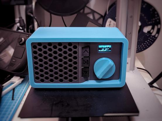front view; completed radio box. Rectangular, blue enclosure with rounded corners, gray font panel with a speaker grill on the left side, three buttons down the center, and volume knob + mini screen on the right side. Entire thing is sitting on a 3D printer plate.