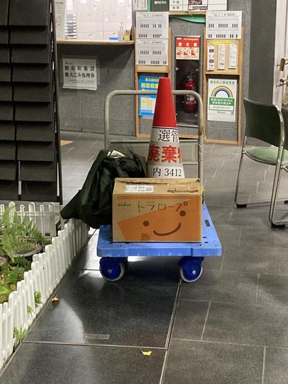 A blue cart with a cardboard box and a green jacket, topped by a red traffic cone with white text. In the background, there are various signs and a fire extinguisher. The setting appears to be an indoor area with tiled flooring and a greenery bed.