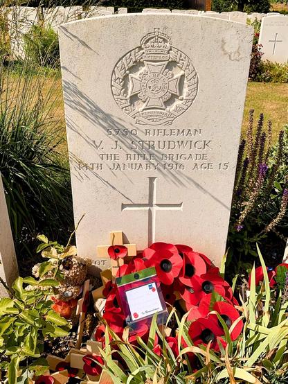 This image shows a war memorial gravestone, honoring a soldier named V. J. Strudwick, who was a rifleman in the Rifle Brigade. He died on January 14, 1916, at the age of 15. The gravestone features a carved emblem with inscriptions and symbols related to the brigade, including “The Prince Consort’s Own.” It is surrounded by red poppies, a common symbol of remembrance for soldiers who have died in war, along with other small remembrance items such as crosses and a poppy wreath. The presence of a small tag and other personal tributes suggests that people continue to visit and honor the memory of this young soldier.
