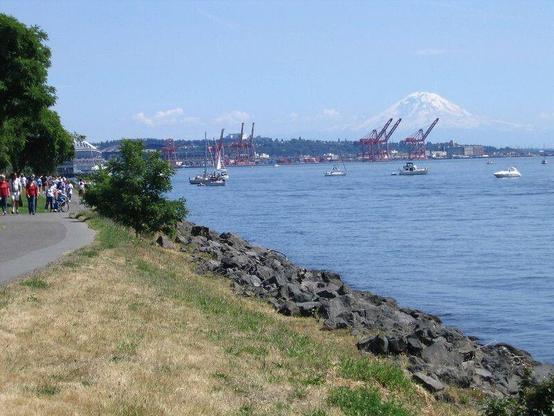 On a gorgeous summer day, people walk along a waterfront path on the left side of the image as boats float in water on the right side of the image, while industrial cranes rise above the port in the distance, and Mt. Rainier  rises over it all in the distance.