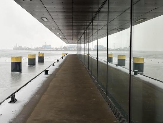 Gebäude mit überdachtem Gang am Alten Hafen in Wismar. Am Rand des Ganges liegt ein Streifen Schnee. Rechts im Bild die Fensterfassade des Gebäudes; hierin spiegeln sich die drei links im Bild sichtbaren schwarz-gelben Metallsäulen und das sie umgebende Ostsee-Wasser des Hafenbeckens. Am Ende des Ganges erkennt man die Silhouette der Gebäude des Seehafens Wismar; wie der Boden des Ganges und die Überdachung spiegeln sich auch die Gebäudesilhouetten in der Fensterfront auf der rechten Seite des Fotos.
