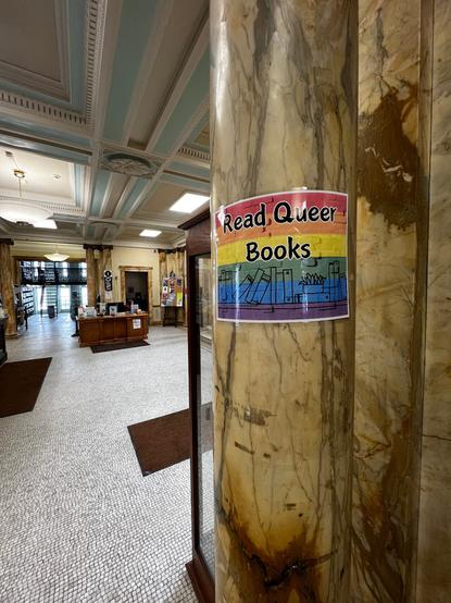 A rainbow-colored sign with a line drawing of books on a shelf, with the title "Read Queer Books" superimposed, is displayed around a marble column in an old city library in Troy, New York.