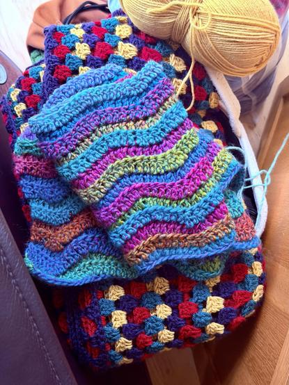 A pair of colourful crochet projects laid out together: a granny‑square blanket made of small red, yellow, blue, and purple squares, and a ripple‑stitch piece with bold wavy stripes in turquoise, pink, green, orange, and more. A ball of yellow yarn sits nearby, showing the blankets are still in progress.