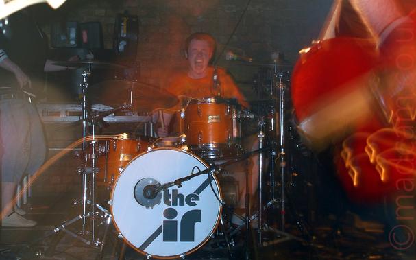 The drummer from a band sitting behind his kit at the back of a stage, and screaming for all he is worth as he plays.
He is wearing an orange t-shirt, which pretty much matches the sides of his drums, and has short dark hair.
The bass drum at the front has a white face, with a diagonal grey strip, overlaid with the grey text "The IR".
Bright lights mounted on metal racks are shining down from the ceiling, in front of a brick wall.
The head of a bass guitar is poking in to frame from the left, with the bottom of a red guitar on the right, with a large expanse of black floor between them.