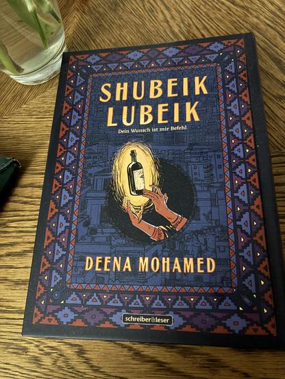 Graphic Novel Shubeik Lubeik, dunkelblau,rot gemustertes Cover mit gezeichneten Händen, die Eine Flasche halten. Auf einem Holztisch.