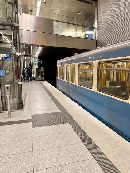 Ein blauer U‑Bahnzug hält an einem modernen, hell beleuchteten unterirdischen Bahnsteig mit Aufzug, taktilen Leitstreifen und Rolltreppe, auf der einige Personen in Richtung Ausgang mit grünen Notausgangsschildern gehen.