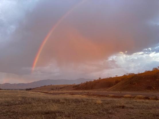 A landscape of grasslands and rolling hills, with barely visible distant mountains. The clouds are lit up orange by the sunset with a short arc of rainbow descending to the ground
